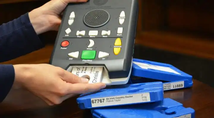 A person inserts a cartridge into a digital audio player for the visually impaired. The device has large, tactile buttons, and blue cases are nearby.