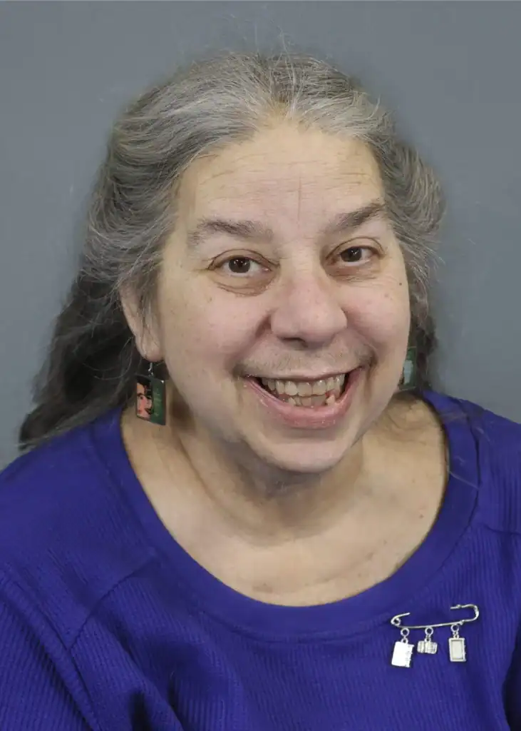 A headshot of board member Mary Nicolini. She is smiling with long gray hair wearing a blue top, set against a plain gray background. Her expression conveys warmth and friendliness.