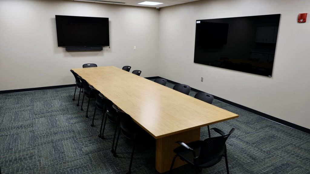 A conference room with a long rectangular wooden table, ten black chairs, a large wall-mounted TV, and a glossy black whiteboard on the wall. The floor is carpeted, and the room is well-lit with overhead lights.