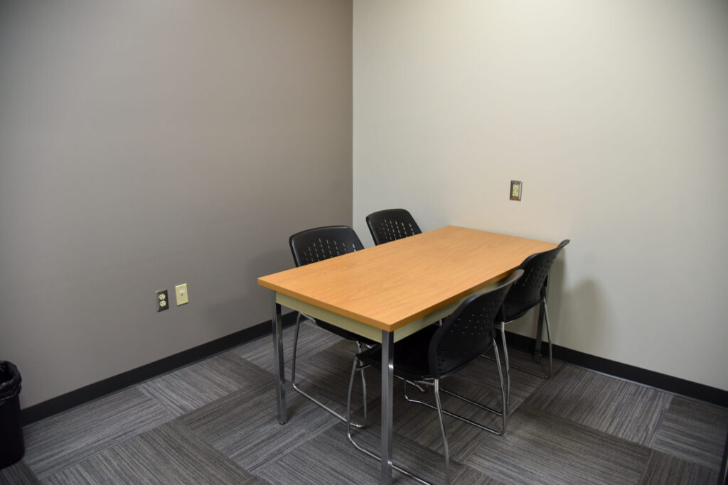 A small meeting room with a rectangular wooden table and four black plastic chairs, set against light gray and white walls with a gray carpeted floor.