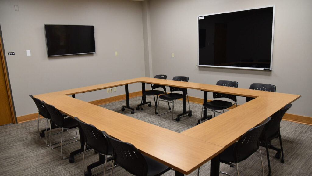 A conference room with a U-shaped table arrangement, black chairs, a wall-mounted flat-screen TV, a large black dry-erase board, and neutral-colored walls and carpet.