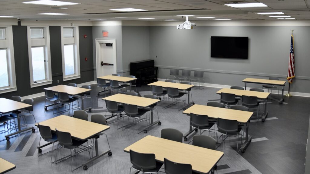 A modern classroom with several rectangular tables and gray chairs arranged in groups, a large wall-mounted TV, an upright piano, and an American flag in the corner. The room is well-lit with large windows.