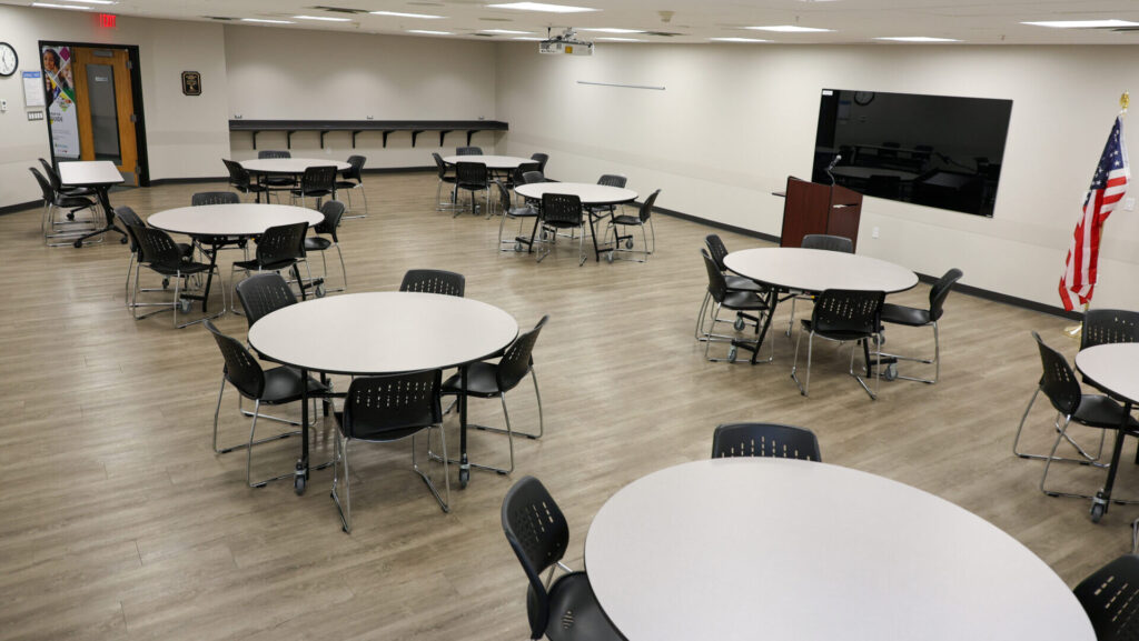 A spacious, well-lit conference room with several round tables and black chairs, a large wall-mounted screen, an American flag, a podium, and light wood flooring. The room appears ready for a meeting or event.