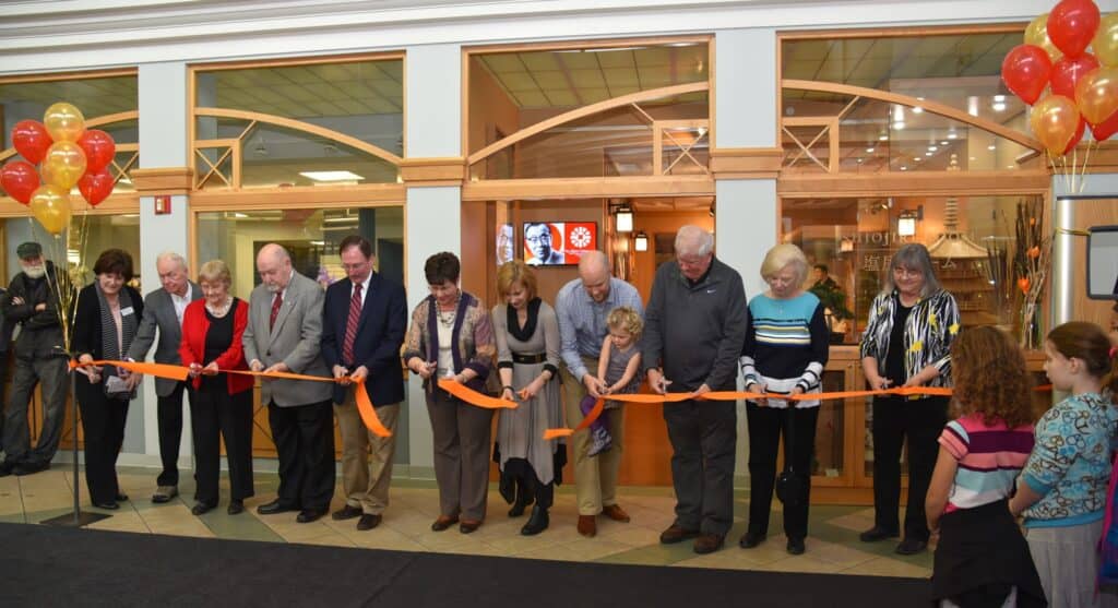 A group of people stand in a line cutting a long orange ribbon at a ribbon-cutting ceremony inside a building. Red and yellow balloons are on the left; two children watch from the right foreground.