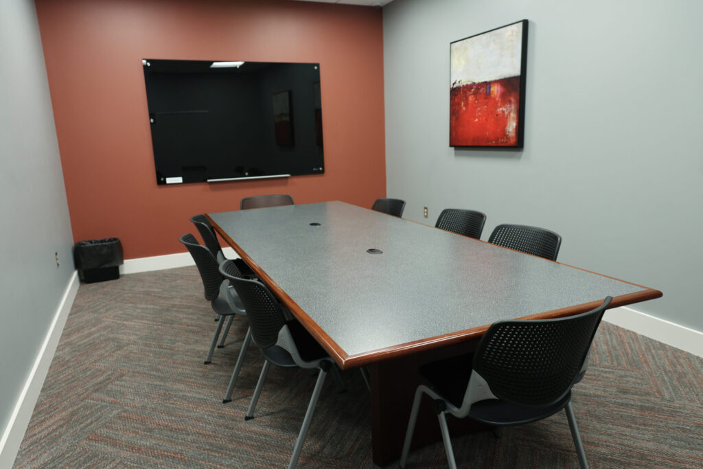 A modern conference room with a long table, eight black chairs, a wall-mounted black whiteboard, a trash bin, and abstract art on a red accent wall. The floor is carpeted with a patterned design.