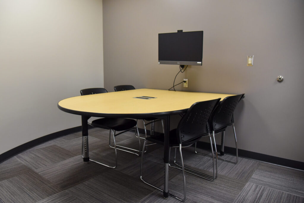 A small conference room with a rectangular table, five black chairs, a wall-mounted monitor, and gray carpeted flooring. The walls are plain and there are no windows or decorations.