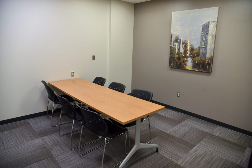 A small conference room with a rectangular wooden table, six black chairs, gray patterned carpet, neutral walls, and a cityscape painting hanging on the wall.