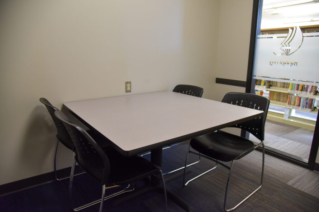 A small meeting room with a square table and four black chairs. The room has a plain wall, carpeted floor, and a window showing bookshelves outside.