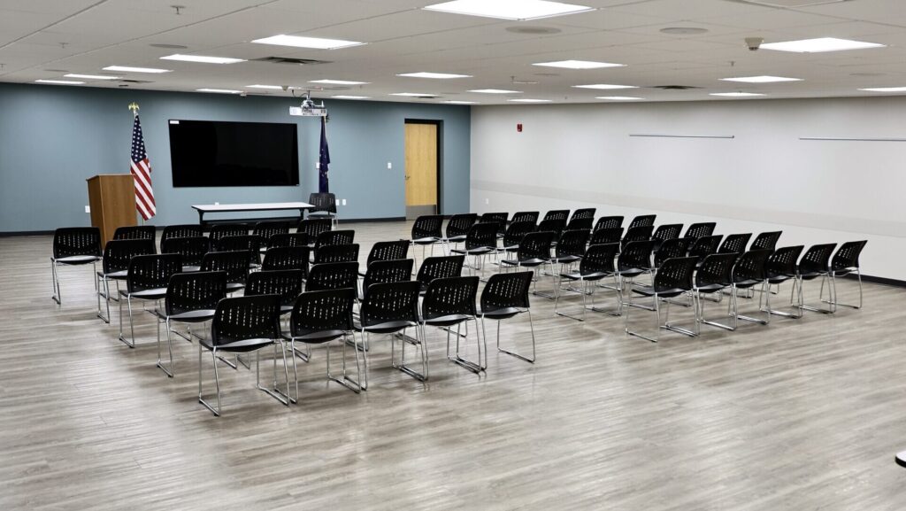 A conference room with rows of black chairs facing a podium, two flags, and a large wall-mounted screen. The room has light wood flooring, white walls, and a modern, organized appearance.