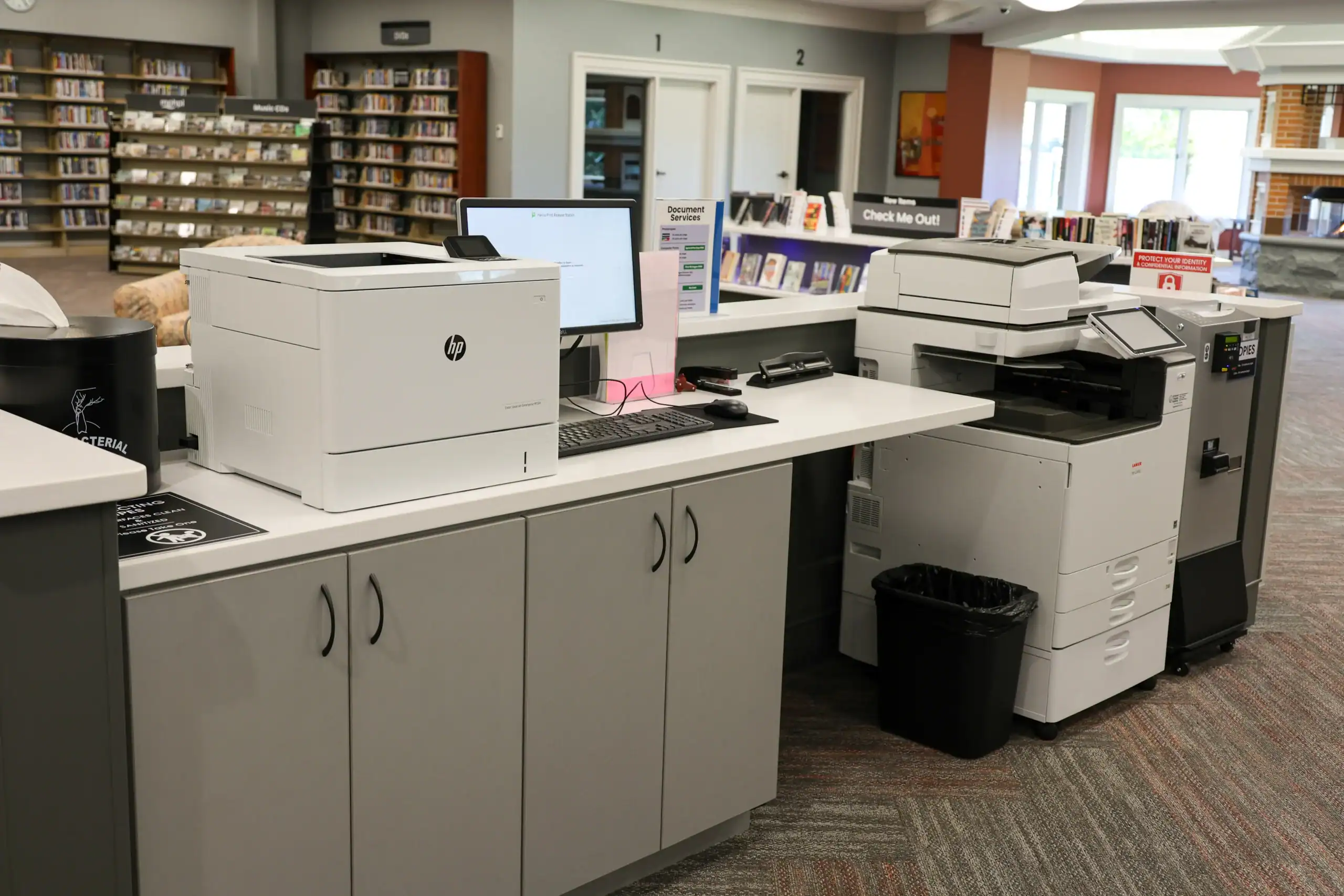 A library service desk with a computer, an HP printer, and a large copier. Bookshelves are visible in the background, along with a Check In/Out sign and a waste bin near the copier.