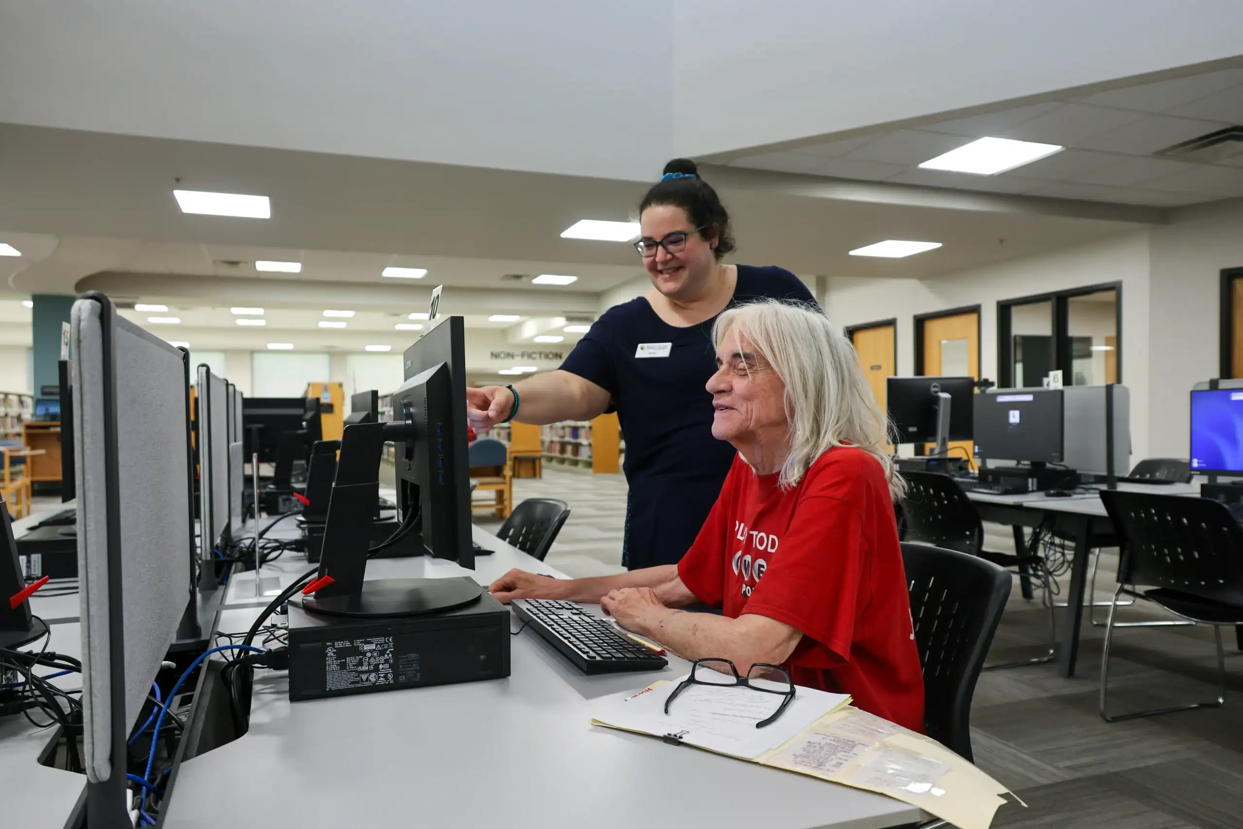 A woman in a red shirt sits at a computer in a library, smiling, as a staff member standing beside her points at the monitor, offering assistance. Other computers and desks are visible in the background.