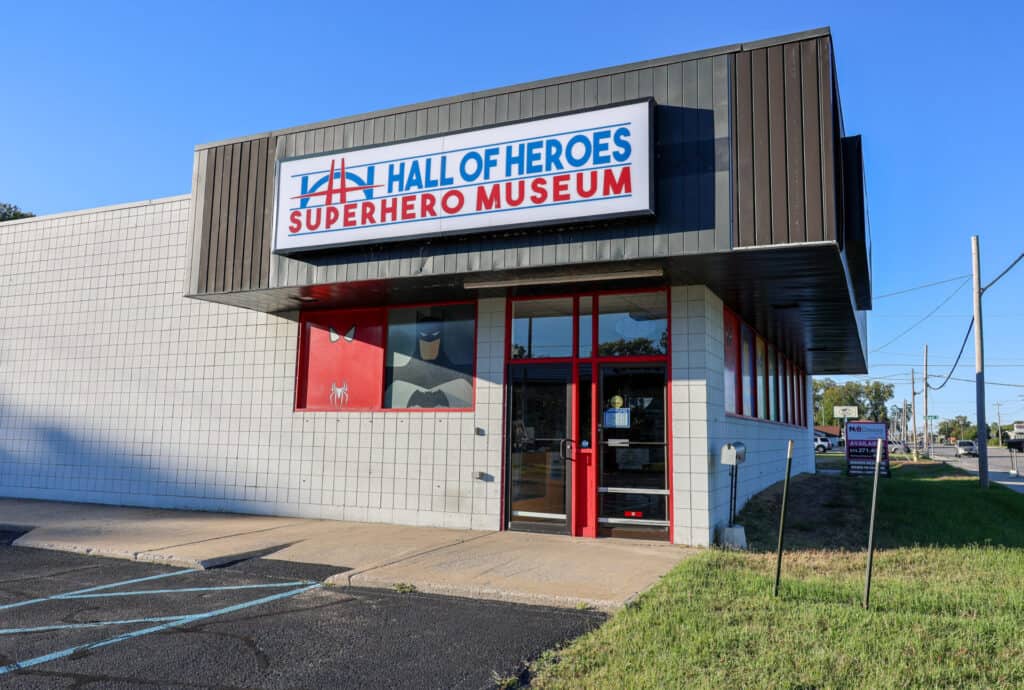 A small building with a sign reading “Hall of Heroes Superhero Museum” above the entrance, which has red-framed glass doors and windows. There is a parking lot and a blue sky in the background.