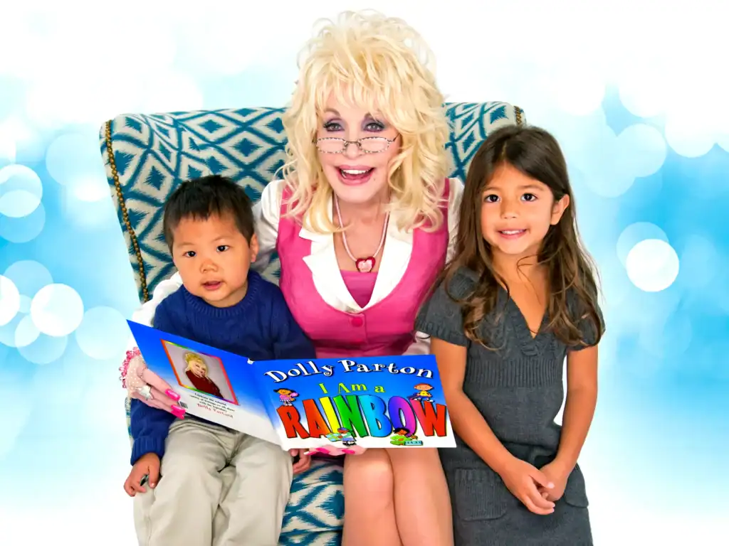 Dolly Parton sits with two children in a chair, as Parton reads a book.